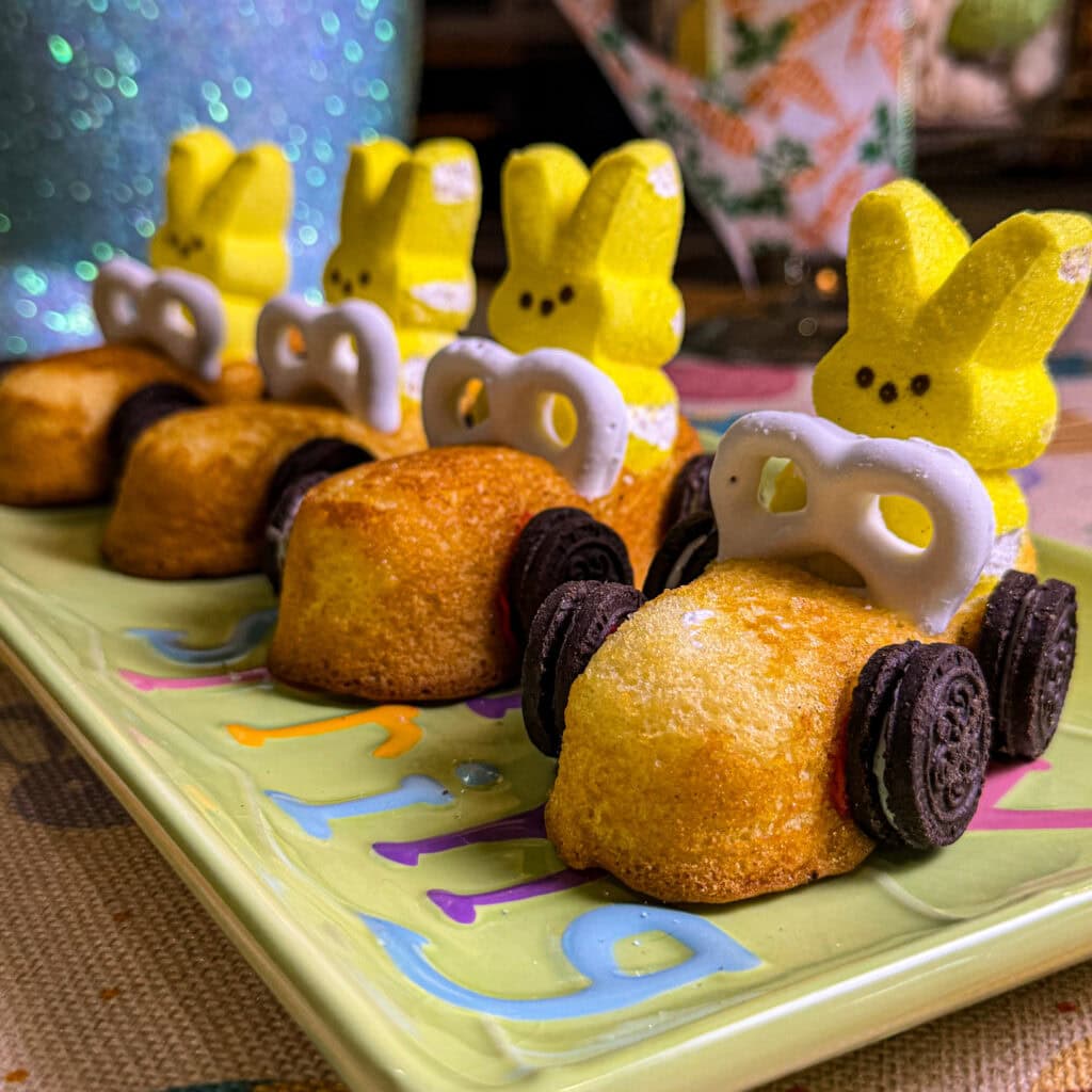 Four Easter treats shaped like race cars, each made from a yellow marshmallow bunny, a small cake, mini chocolate cookies for wheels, and a white pretzel front—perfect alongside your Overnight Hashbrown Breakfast Casserole on a pastel plate.
