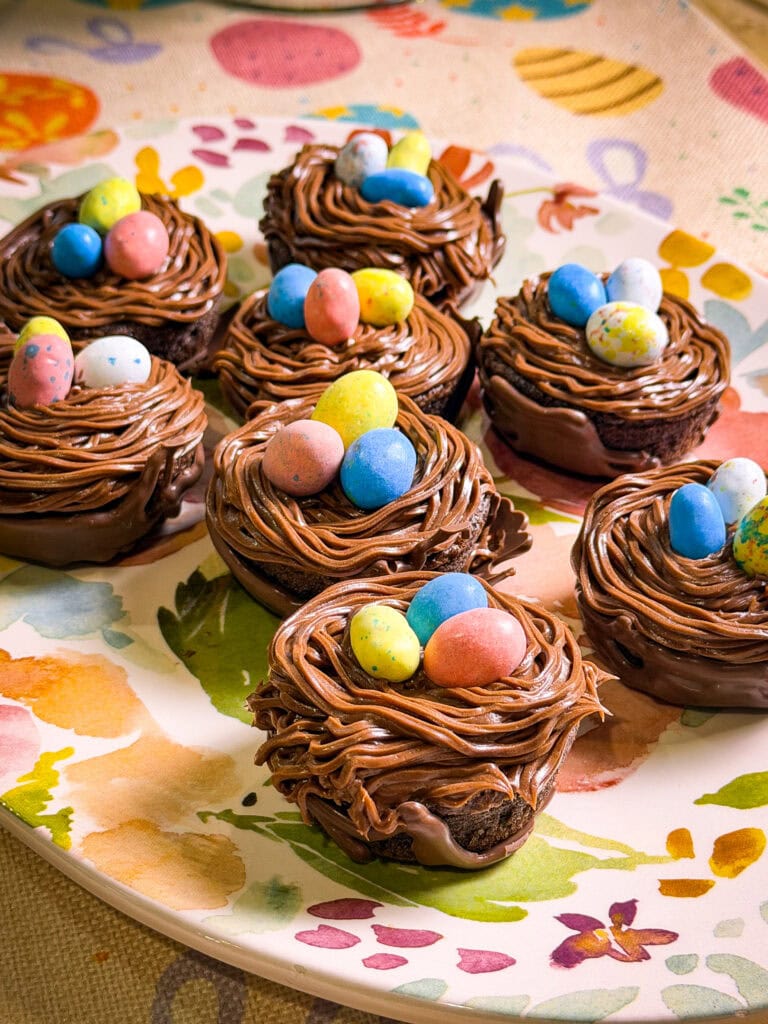 Chocolate cupcakes decorated to look like bird nests with chocolate frosting and colorful candy eggs on top, arranged on a floral-patterned plate—perfect for an Easter or spring-themed table alongside your Overnight Hashbrown Breakfast Casserole.