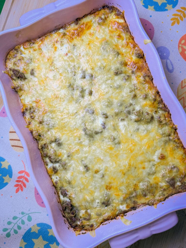 A rectangular casserole dish filled with a baked, golden-brown Overnight Hashbrown Breakfast Casserole sits on a colorful, patterned tablecloth with Easter egg designs. The dish has browned edges and a bubbly cheese topping.