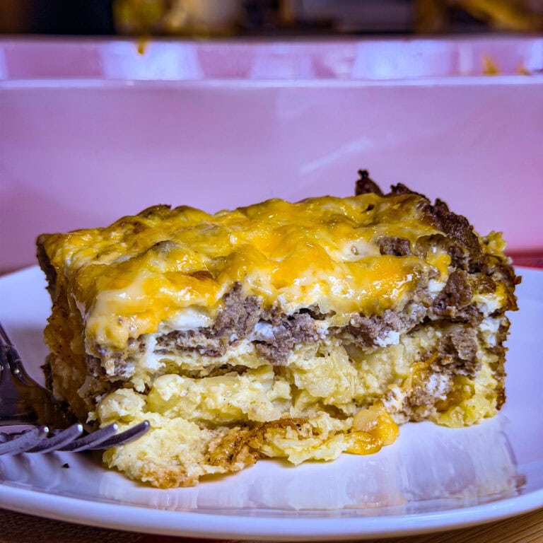 A close-up of a slice of cheesy Overnight Hashbrown Breakfast Casserole with layers of eggs, sausage, and melted cheese on a white plate, with a fork beside it. The background shows the casserole dish.