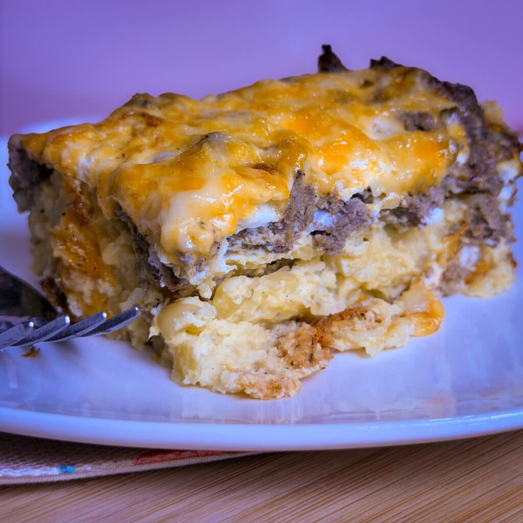 A close-up of an Overnight Hashbrown Breakfast Casserole with layers of eggs, sausage, hashbrowns, and cheese on a white plate, with a fork beside the serving.