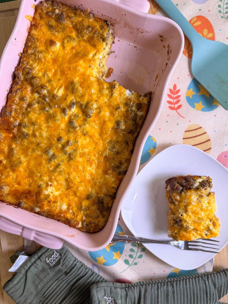 A baked cheesy Overnight Hashbrown Breakfast Casserole sits in a pink dish with a serving removed. The slice is on a white plate with a fork, alongside an Easter-themed napkin and green oven mitts.