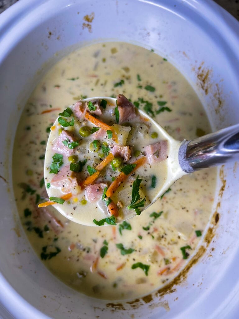 creamy soup of ham, carrots, potatoes and peas in a soup ladle scooping from a crockpot