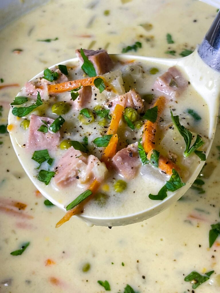 creamy soup of ham, carrots, potatoes and peas in a soup ladle scooping from a crockpot