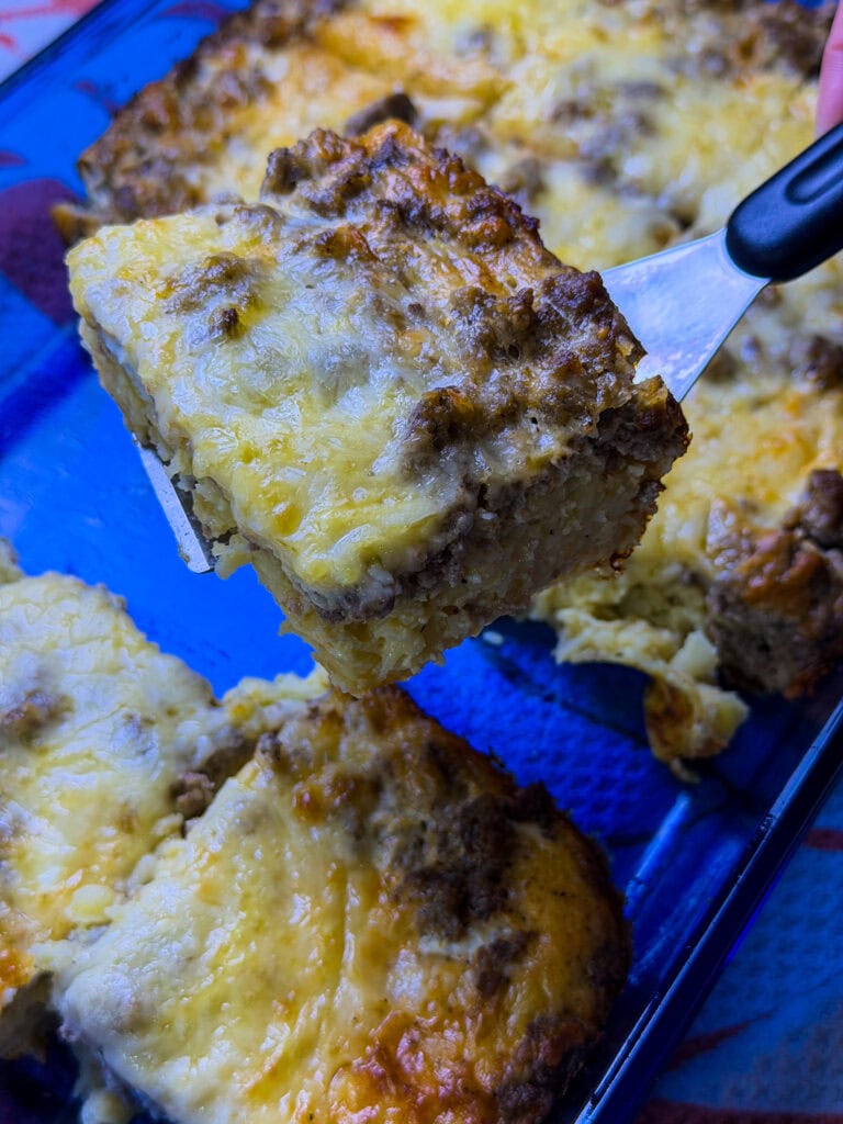 A slice of breakfast casserole made with hashbrowns, sausage, eggs, and cheese on a blue plate with the blue casserole serving dish in the background