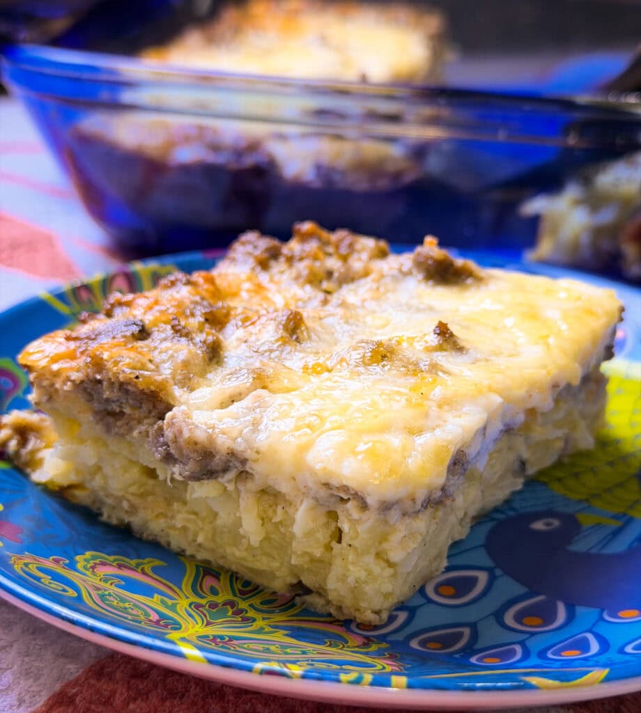 A slice of breakfast casserole made with hashbrowns, sausage, eggs, and cheese on a blue plate with the blue casserole serving dish in the background