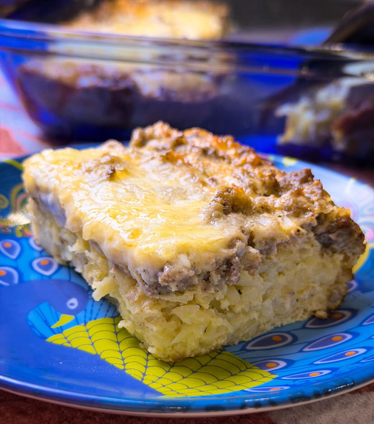 A slice of breakfast casserole made with hashbrowns, sausage, eggs, and cheese on a blue plate with the blue casserole serving dish in the background