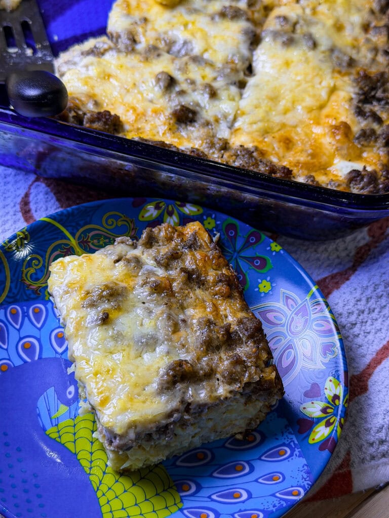 A slice of breakfast casserole made with hashbrowns, sausage, eggs, and cheese on a blue plate with the blue casserole serving dish in the background