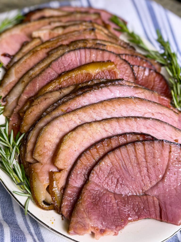 spiral slices of ham made with cherry coke glaze laid out on a white serving platter with sprigs of rosemary
