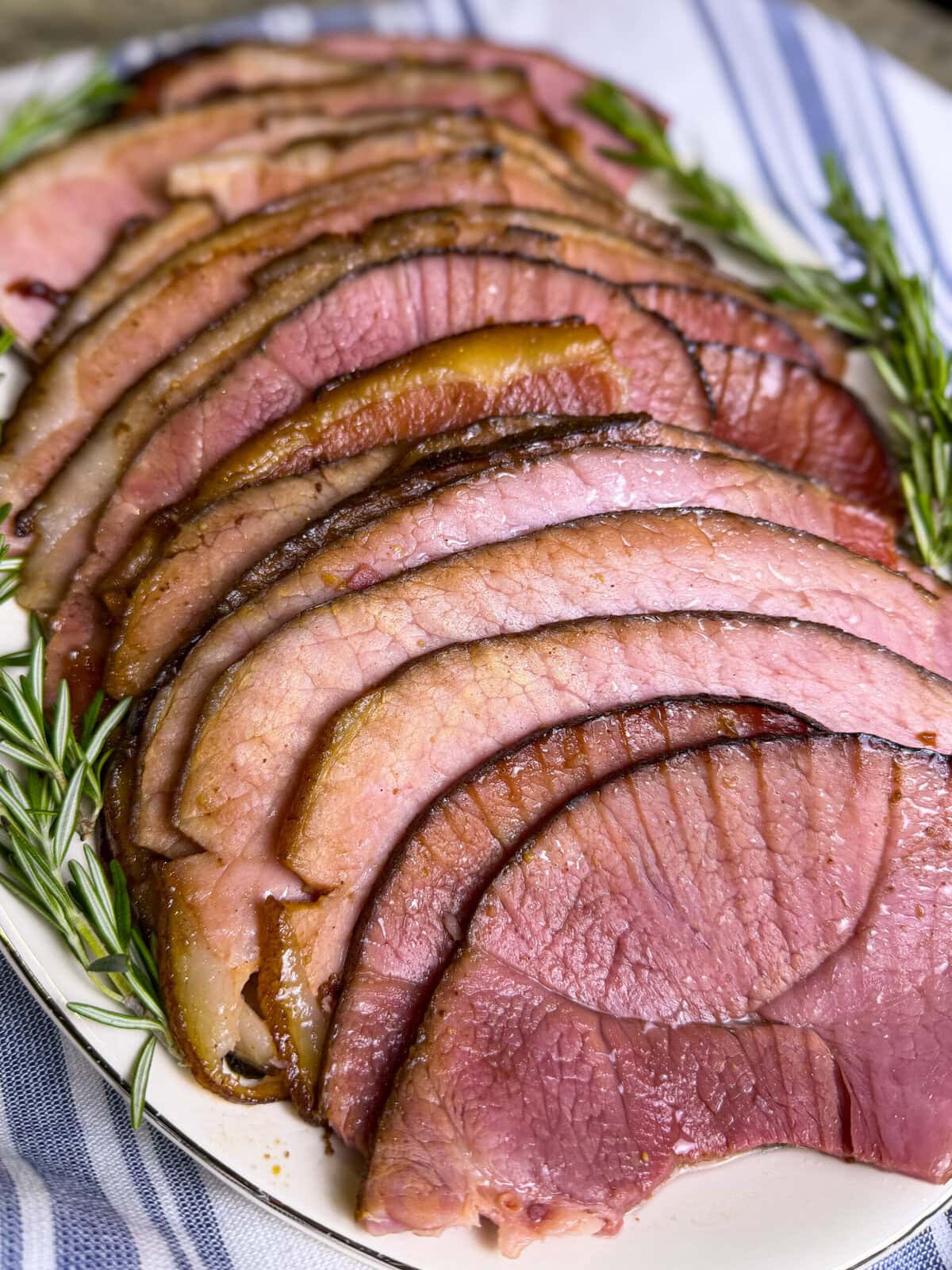 spiral slices of ham made with cherry coke glaze laid out on a white serving platter with sprigs of rosemary
