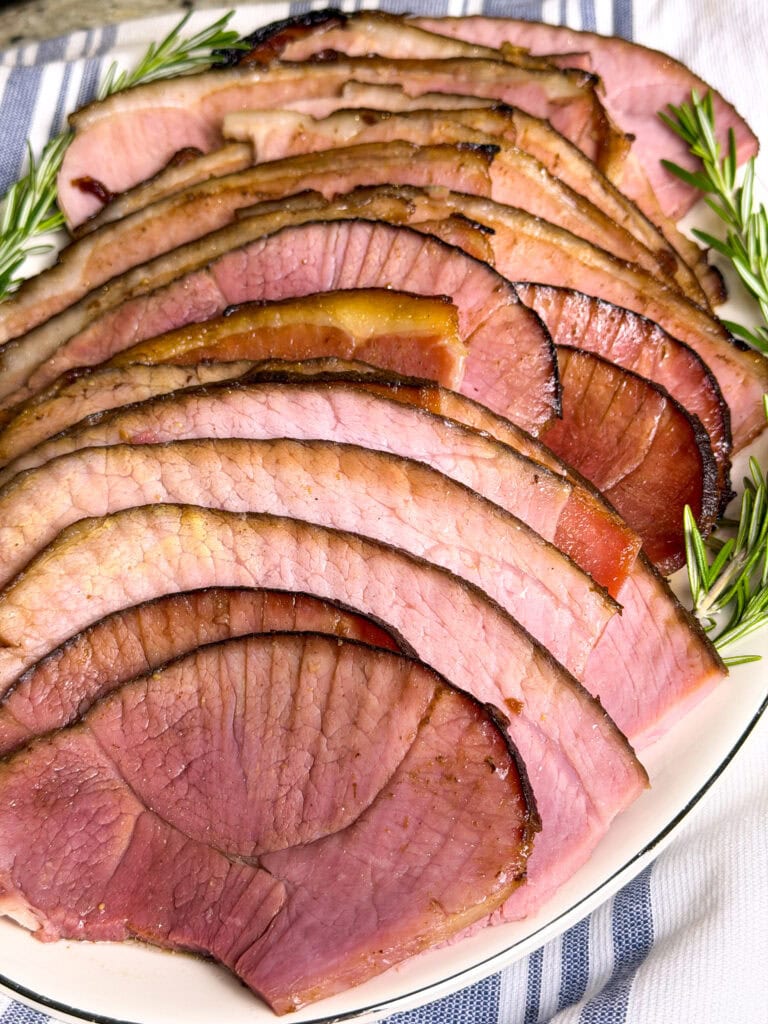 spiral slices of ham made with cherry coke glaze laid out on a white serving platter with sprigs of rosemary