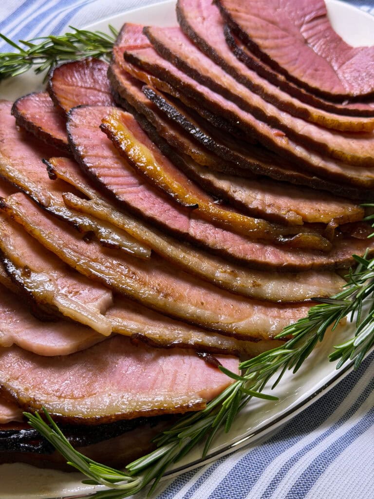 spiral slices of ham made with cherry coke glaze laid out on a white serving platter with sprigs of rosemary