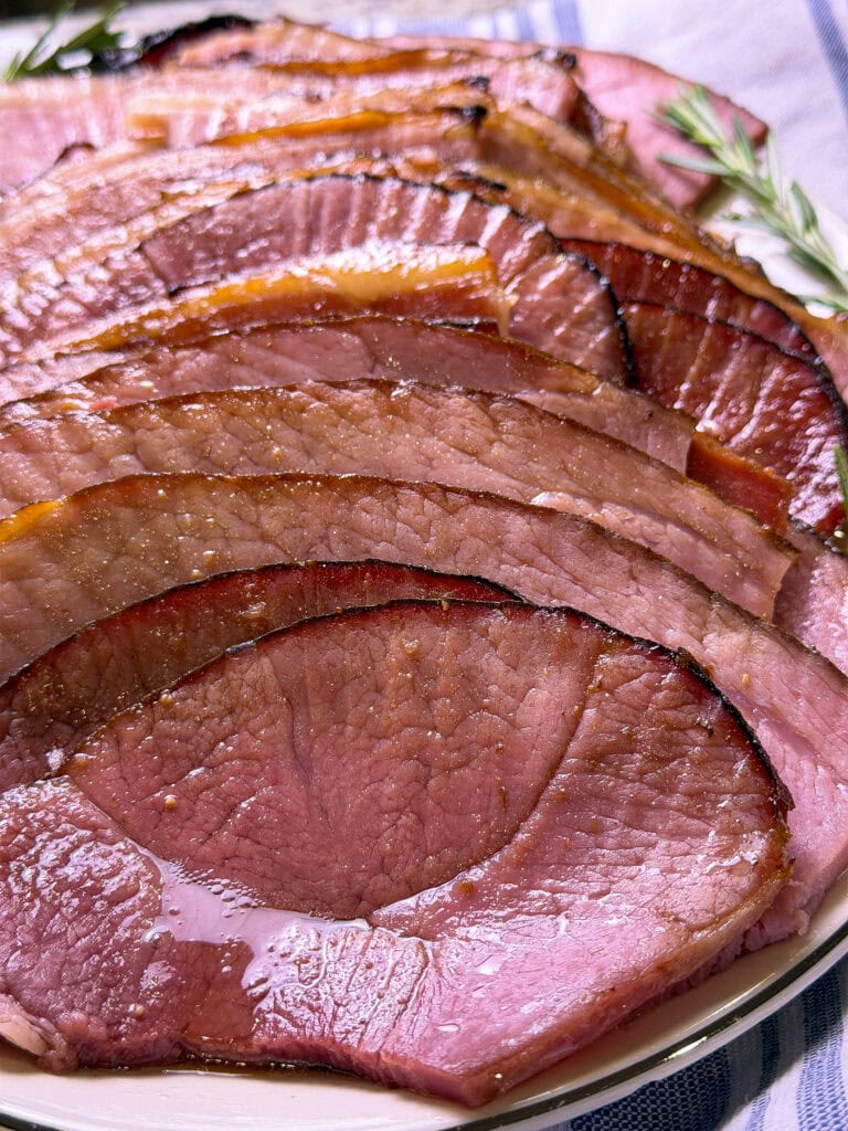 spiral slices of ham made with cherry coke glaze laid out on a white serving platter with sprigs of rosemary