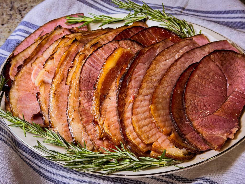 spiral slices of ham made with cherry coke glaze laid out on a white serving platter with sprigs of rosemary