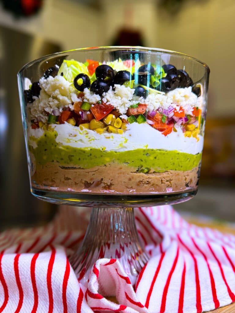 seven layer dip in a glass trifle dish, layered from bottom to top: refried beans, guacamole. sour cream, Pico de Gallow, roasted corn, queso fresco, black olives and shredded lettuce