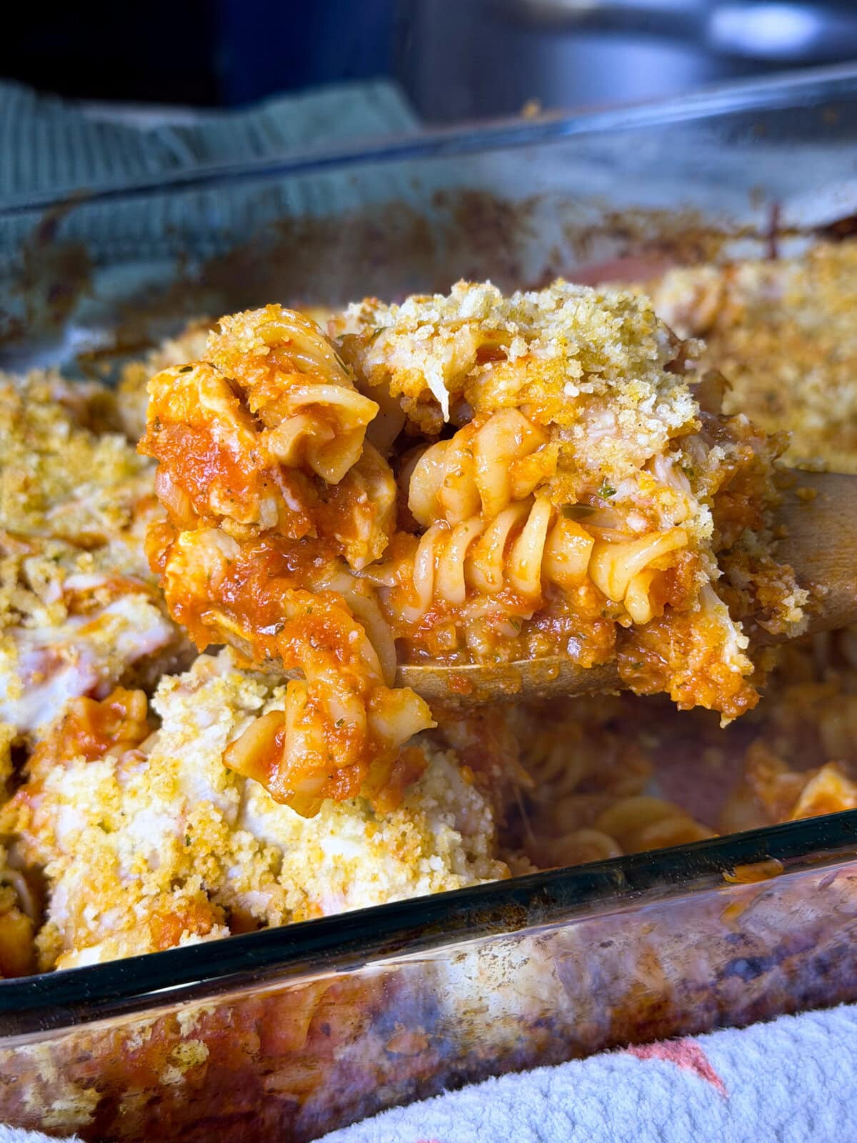 A golden, cheesy baked chicken Parmesan casserole in a large white dish, topped with crispy seasoned breadcrumbs and melted mozzarella, with tender pasta and pieces of chicken visible underneath.