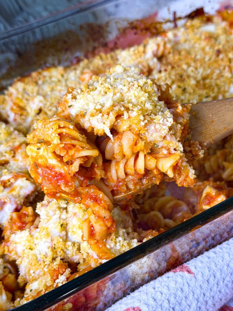 A golden, cheesy baked chicken Parmesan casserole in a large white dish, topped with crispy seasoned breadcrumbs and melted mozzarella, with tender pasta and pieces of chicken visible underneath.