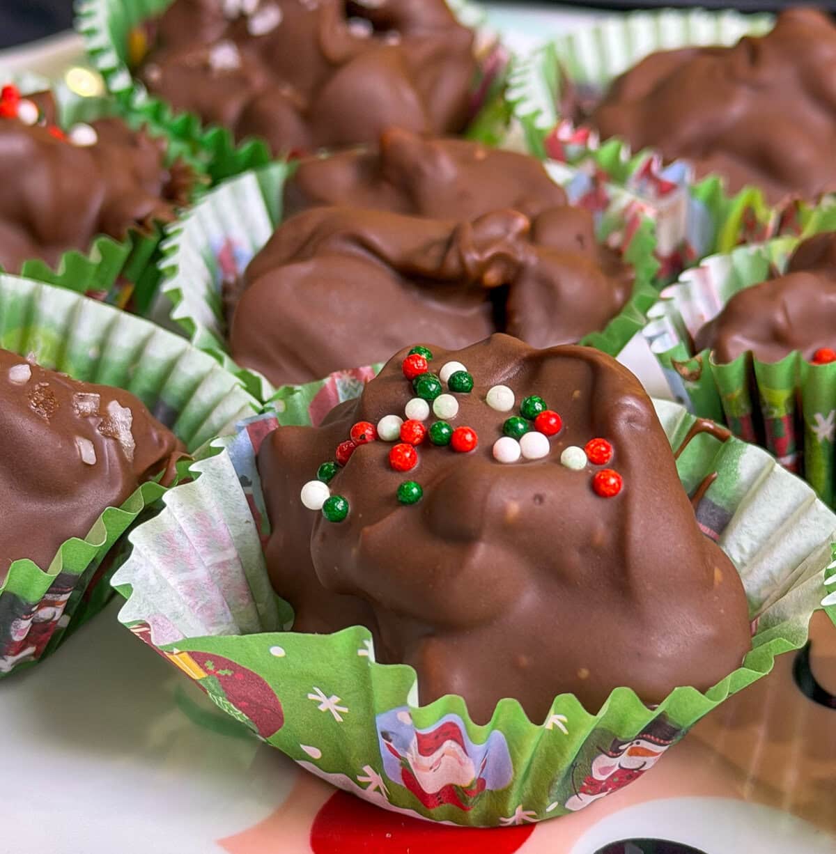 chocolate covered mixed nuts with pretzels scooped into a green mini muffin liner with red, green, and white sprinkles all made in the slow cooker