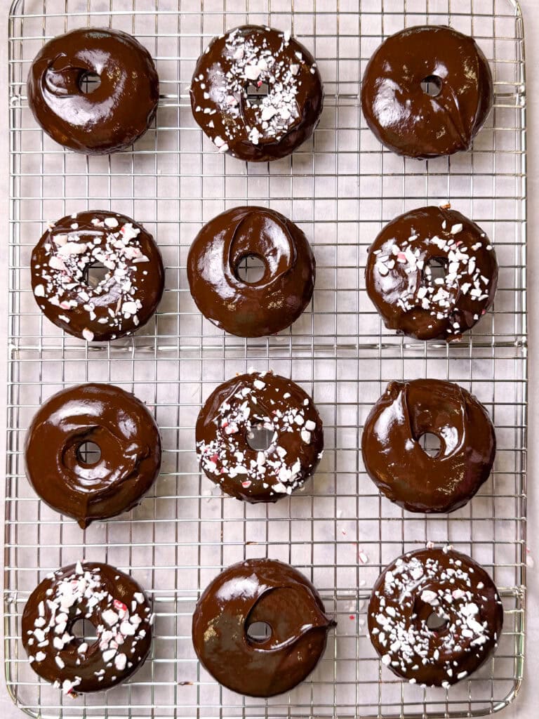 Baked chocolate peppermint donuts dipped in glossy chocolate ganache and sprinkled with crushed candy canes on a cooling rack.
