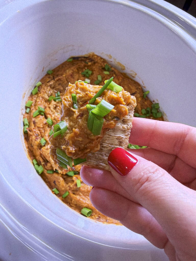 shredded chicken cream cheese sweet chili sauce dip in a white crock pot topped with sliced green onions with a scoop on a Triscuit cracker