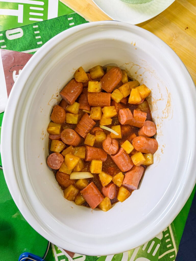 kielbasa cut hasselback style in a sweet and savory BBQ brown sugar sauce with onion slices and pineapple chunks in a white crock pot