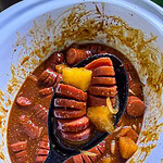 kielbasa cut hasselback style in a sweet and savory BBQ brown sugar sauce with onion slices and pineapple chunks in a black spoon