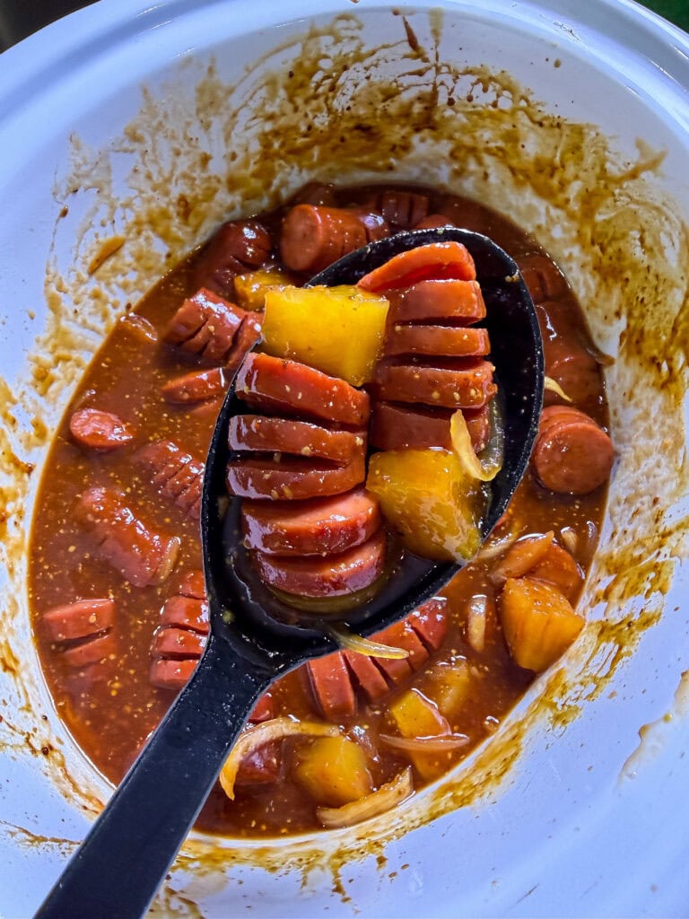 kielbasa cut hasselback style in a sweet and savory BBQ brown sugar sauce with onion slices and pineapple chunks in a black spoon