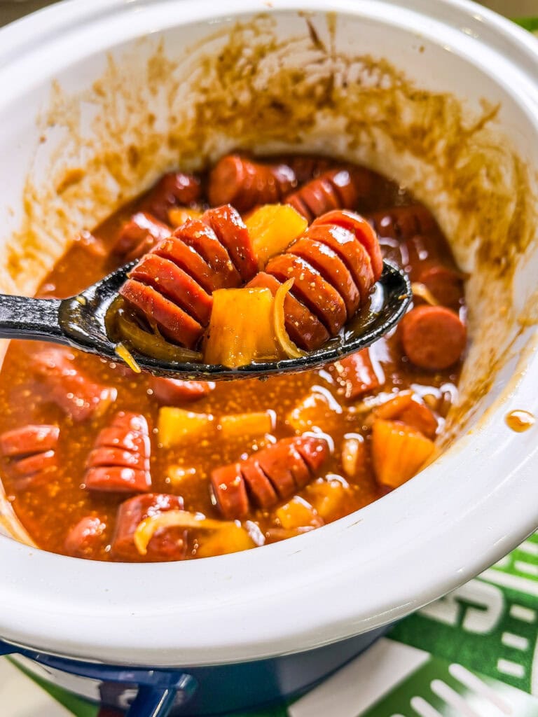 kielbasa cut hasselback style in a sweet and savory BBQ brown sugar sauce with onion slices and pineapple chunks in a black spoon