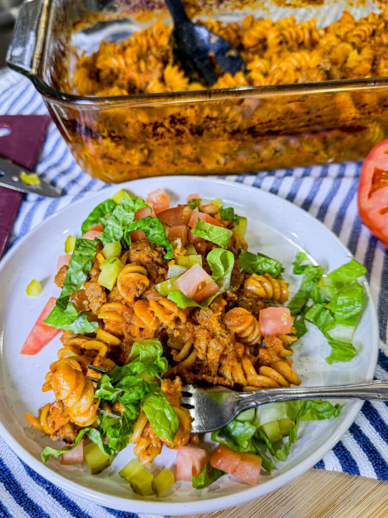 One-dish baked cheeseburger pasta served on a plate with melted cheddar cheese, diced pickles, shredded lettuce, and chopped tomatoes.