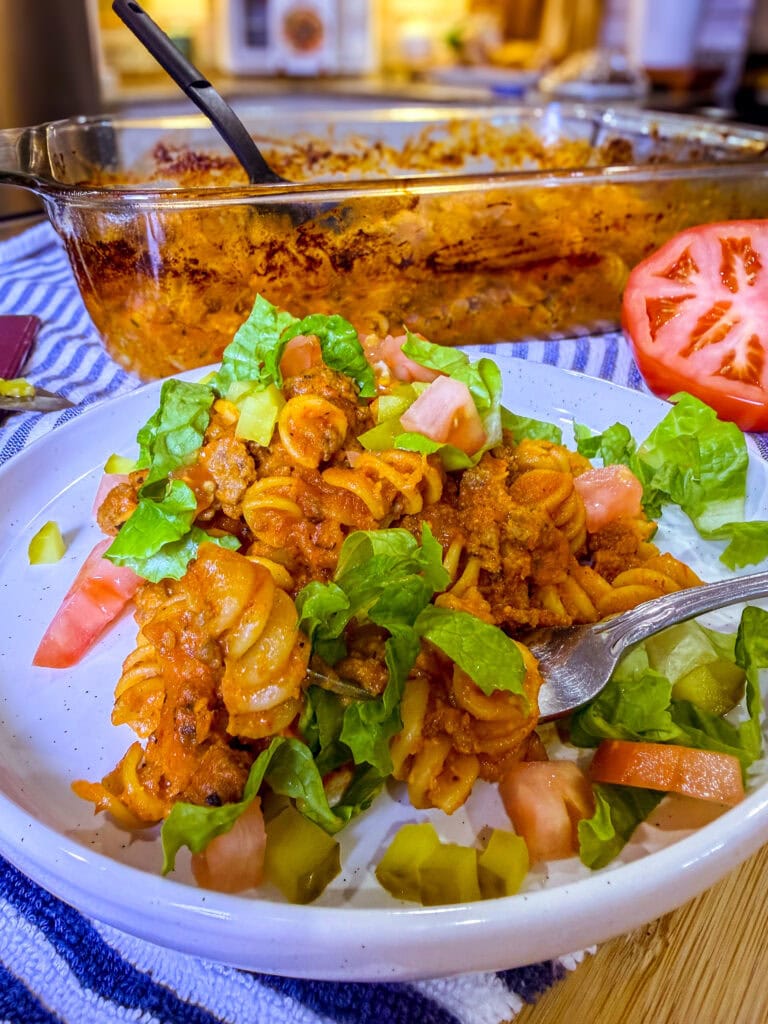One-dish baked cheeseburger pasta served on a plate with melted cheddar cheese, diced pickles, shredded lettuce, and chopped tomatoes.
