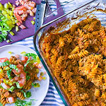 One-dish baked cheeseburger pasta served on a plate with melted cheddar cheese, diced pickles, shredded lettuce, and chopped tomatoes.