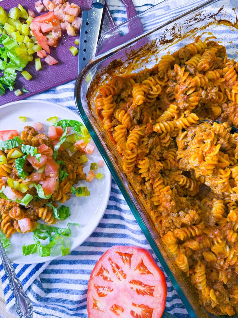 One-dish baked cheeseburger pasta served on a plate with melted cheddar cheese, diced pickles, shredded lettuce, and chopped tomatoes.