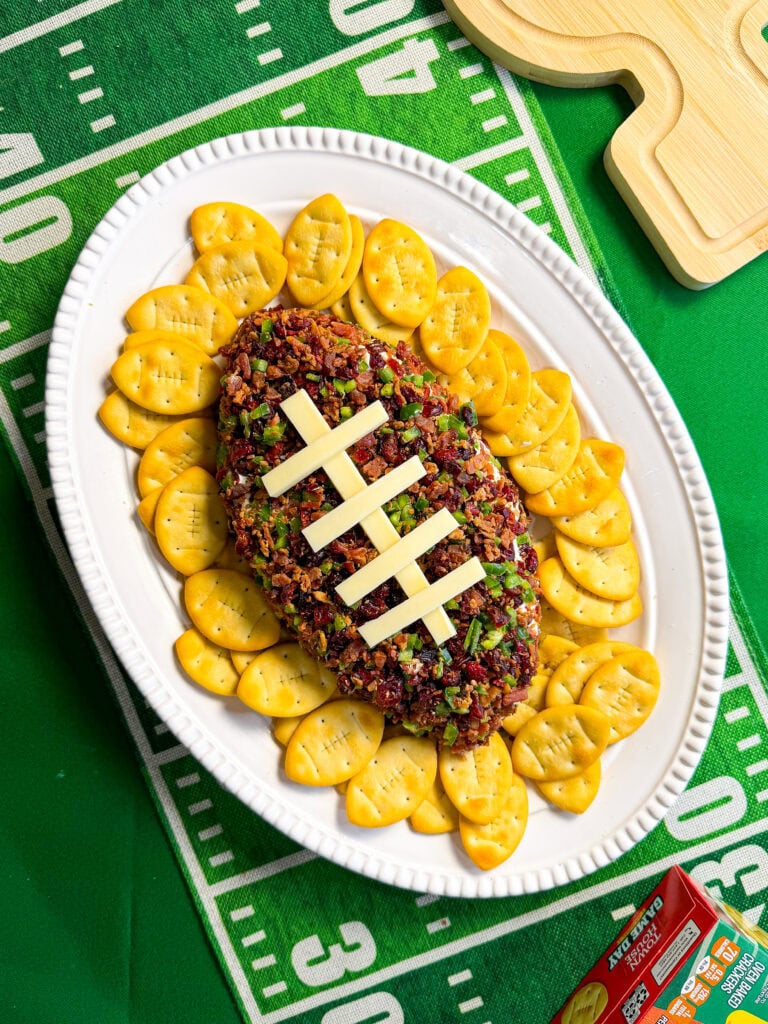 Jalapeño Popper Football Dip shaped like a football with cheese laces, bacon, cranberries, and jalapeños on a game day snack table