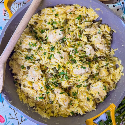 creamy chicken piccata with orzo being served from a yellow skillet with a wooden spoon resting on the skillet