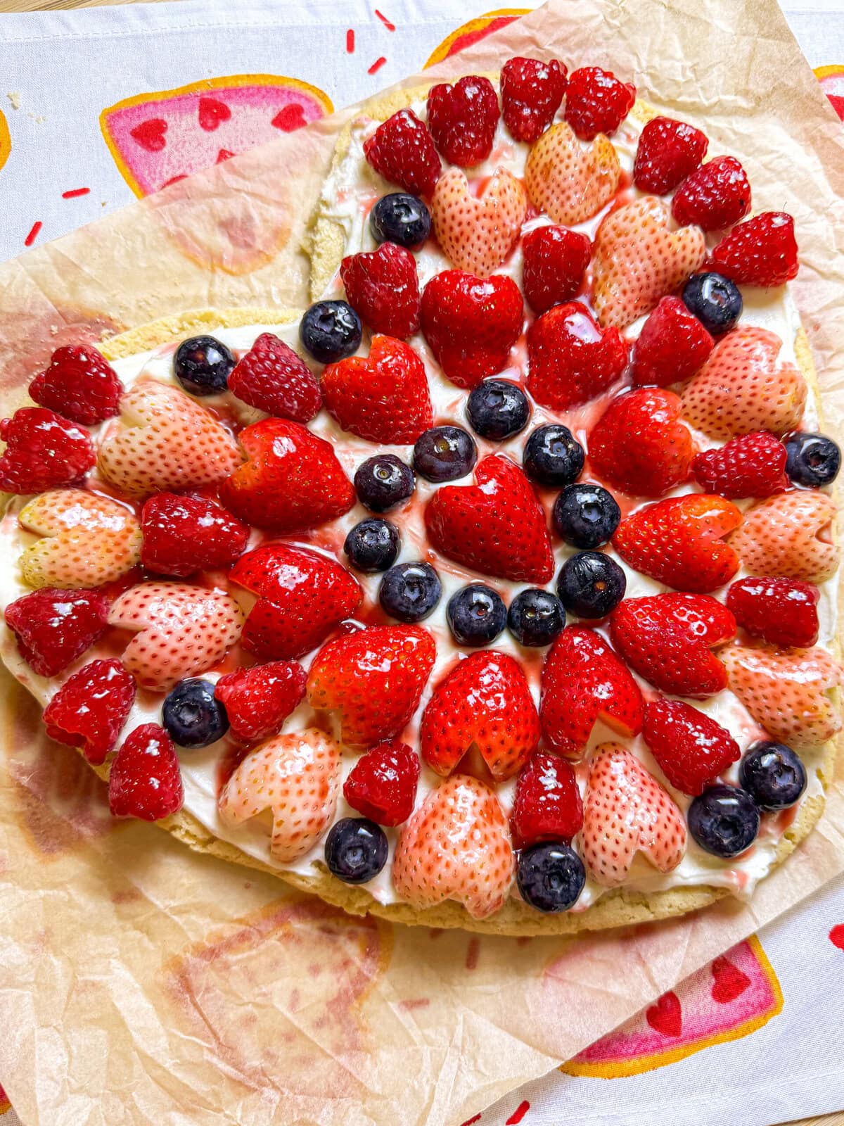 Heart shaped sugar cookie fruit pizza topped with cream cheese frosting, strawberries, blueberries, raspberries, and a glossy jam glaze for Valentine’s Day.