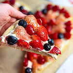 a close up of a slice from a Heart shaped sugar cookie fruit pizza topped with cream cheese frosting, strawberries, blueberries, raspberries, and a glossy jam glaze for Valentine’s Day.
