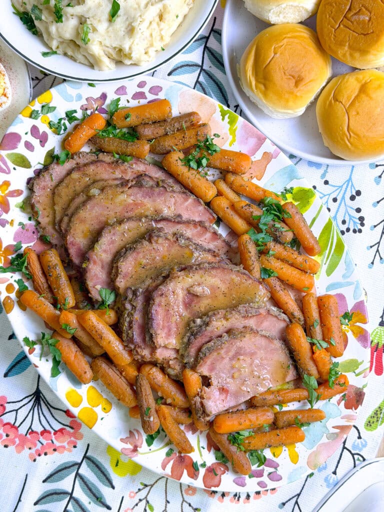 glazed ham slices on a flower platter surrounded by baby carrots with dinner rolls and mashed potatoes in the background