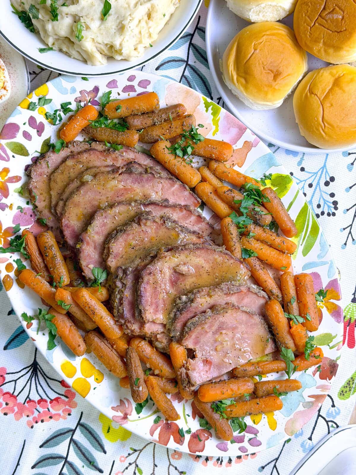 glazed ham slices on a flower platter surrounded by baby carrots with dinner rolls and mashed potatoes in the background