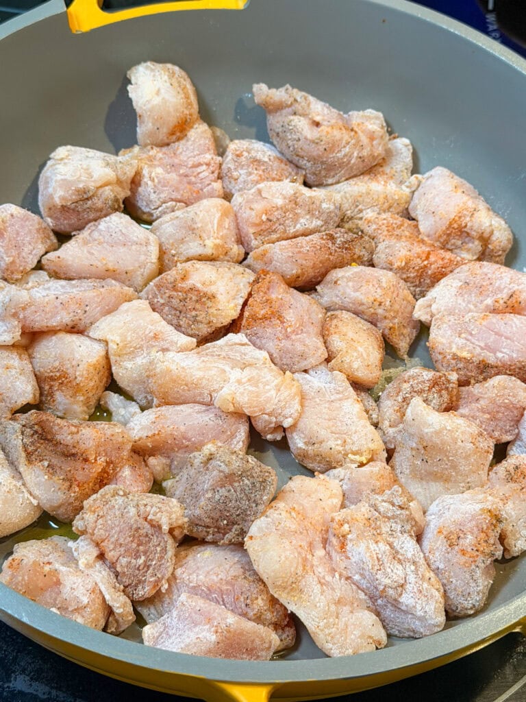 Raw, seasoned chicken pieces coated with flour are spread out evenly in a gray frying pan with a yellow handle, ready to be cooked for a delicious One Pot Chicken Piccata with Orzo.