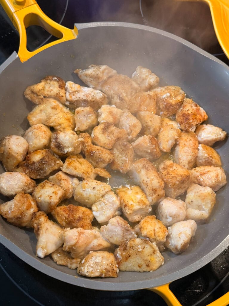 Chunks of seasoned chicken cooking in a gray nonstick skillet with yellow handles, surrounded by steam rising from the pan, create the mouthwatering beginning of this One Pot Chicken Piccata with Orzo.