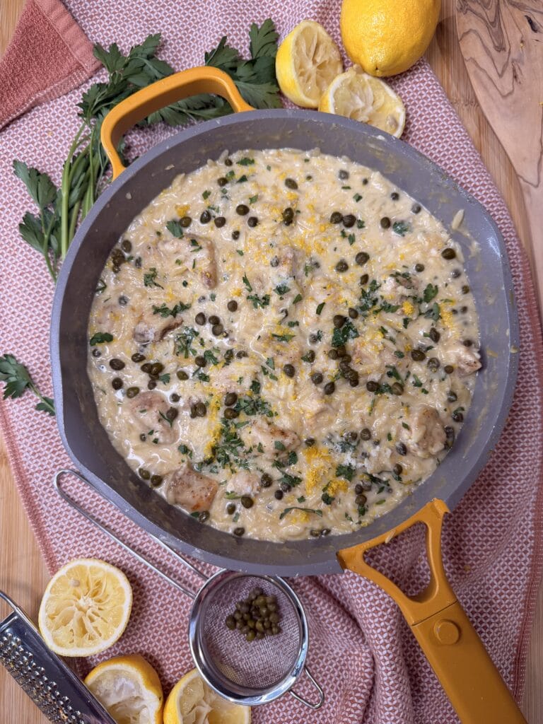 A skillet of creamy One Pot Chicken Piccata with Orzo, garnished with parsley and capers, sits on a wooden surface alongside sliced lemons, parsley, a grater, and a strainer.