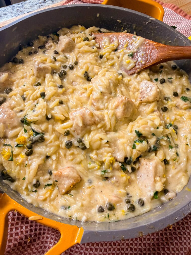A skillet filled with creamy orzo pasta, chunks of chicken, capers, herbs, and a wooden spoon resting on the edge captures the essence of One Pot Chicken Piccata with Orzo.