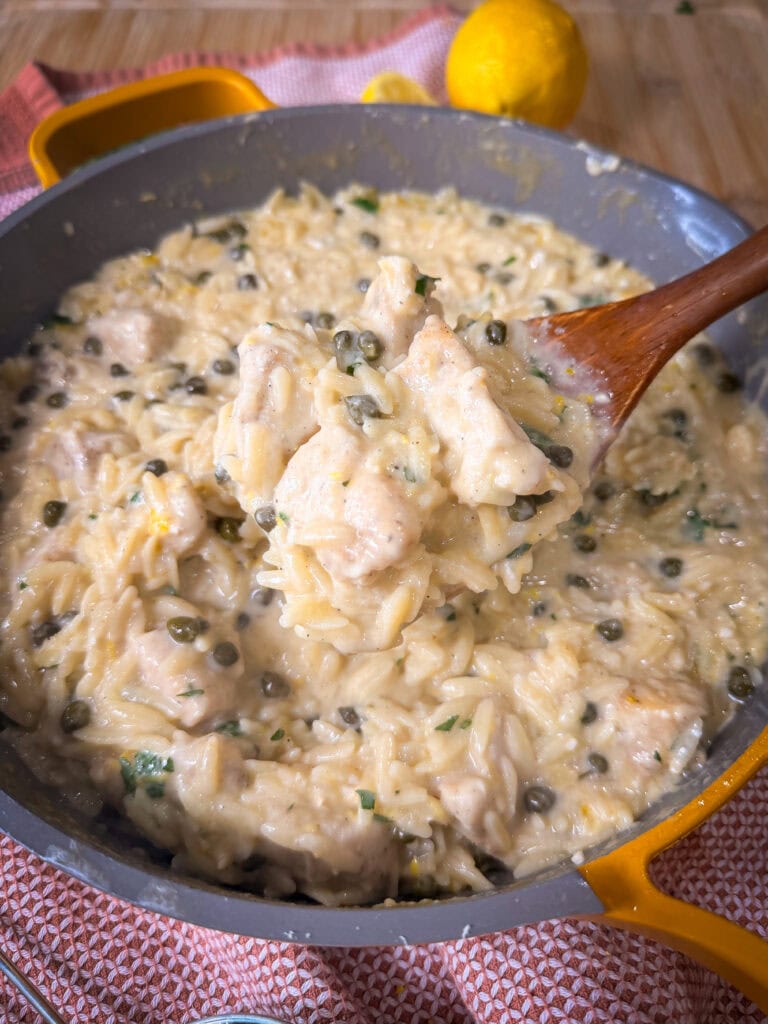Creamy One Pot Chicken Piccata with Orzo, capers, and herbs in a gray skillet, with a wooden spoon lifting a portion. A lemon and red cloth are visible in the background.