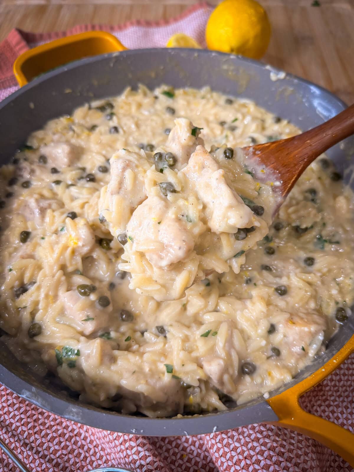 Creamy One Pot Chicken Piccata with Orzo, capers, and herbs in a gray skillet, with a wooden spoon lifting a portion. A lemon and red cloth are visible in the background.