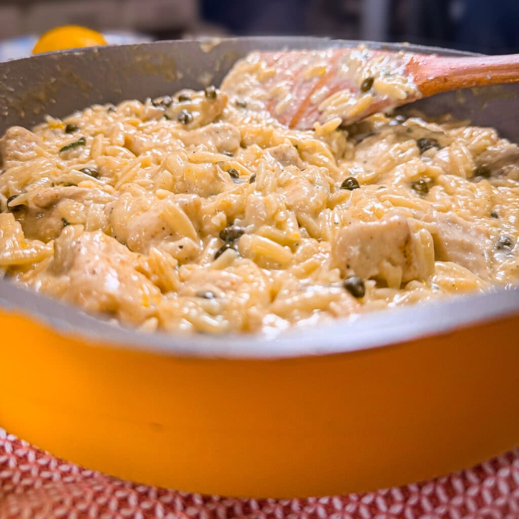 Creamy orzo pasta with chicken and herbs, reminiscent of a One Pot Chicken Piccata with Orzo, being stirred in a yellow pan with a wooden spoon, set on a red and white textured mat.