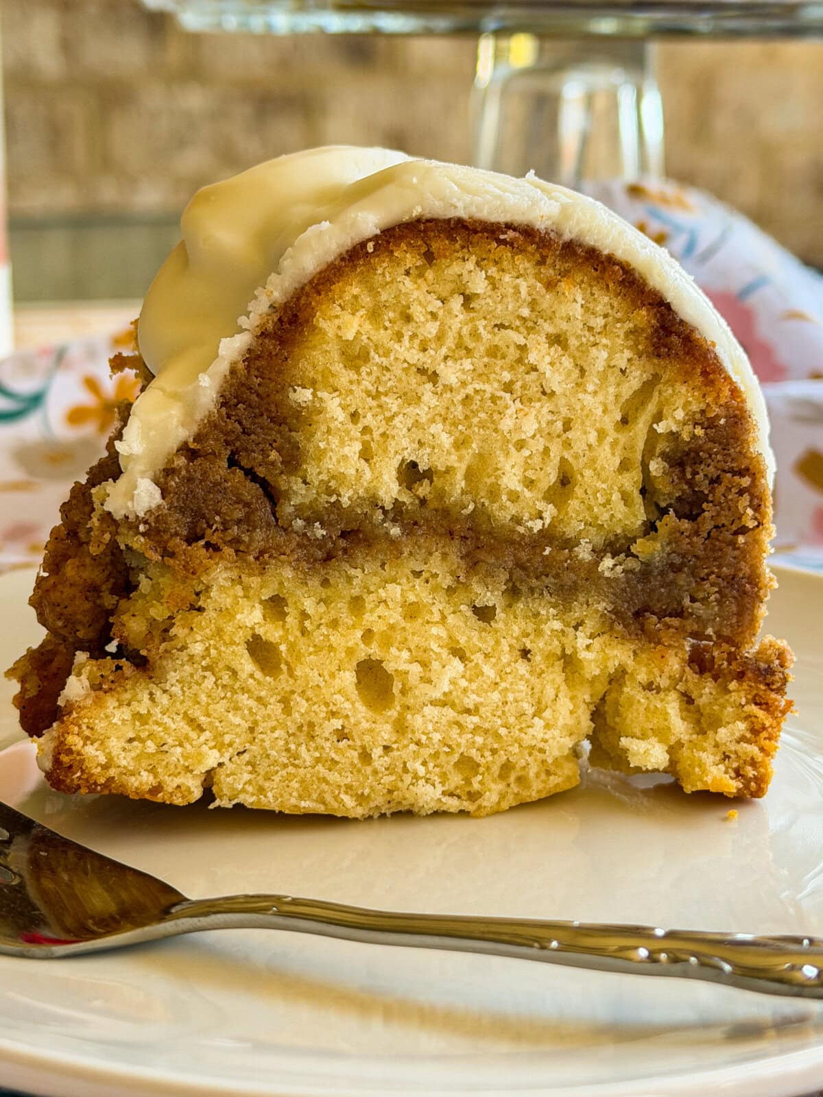 A close-up of a slice of bundt cake with a cinnamon swirl and creamy white icing on top, served on a white plate with a silver fork beside it. The background is softly blurred.