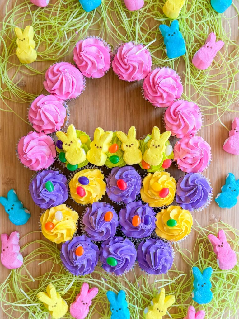 Cupcakes with pink, yellow, and purple frosting are arranged in the shape of an Easter basket, decorated with marshmallow bunny candies and jelly beans—one of the easy Easter treat ideas—on a wooden surface with fake grass and more bunny candies surrounding it.