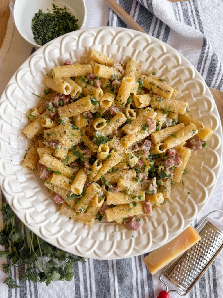 A plate of rigatoni pasta mixed with diced meat, sprinkled with chopped parsley and grated cheese, sits on a white textured dish next to a bowl of herbs, a grater, a wedge of cheese, and fresh parsley.