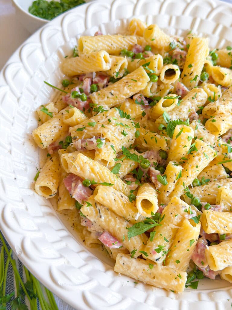A close-up of a white bowl filled with creamy rigatoni pasta, diced ham, herbs, and green garnish, set on a light-colored table.