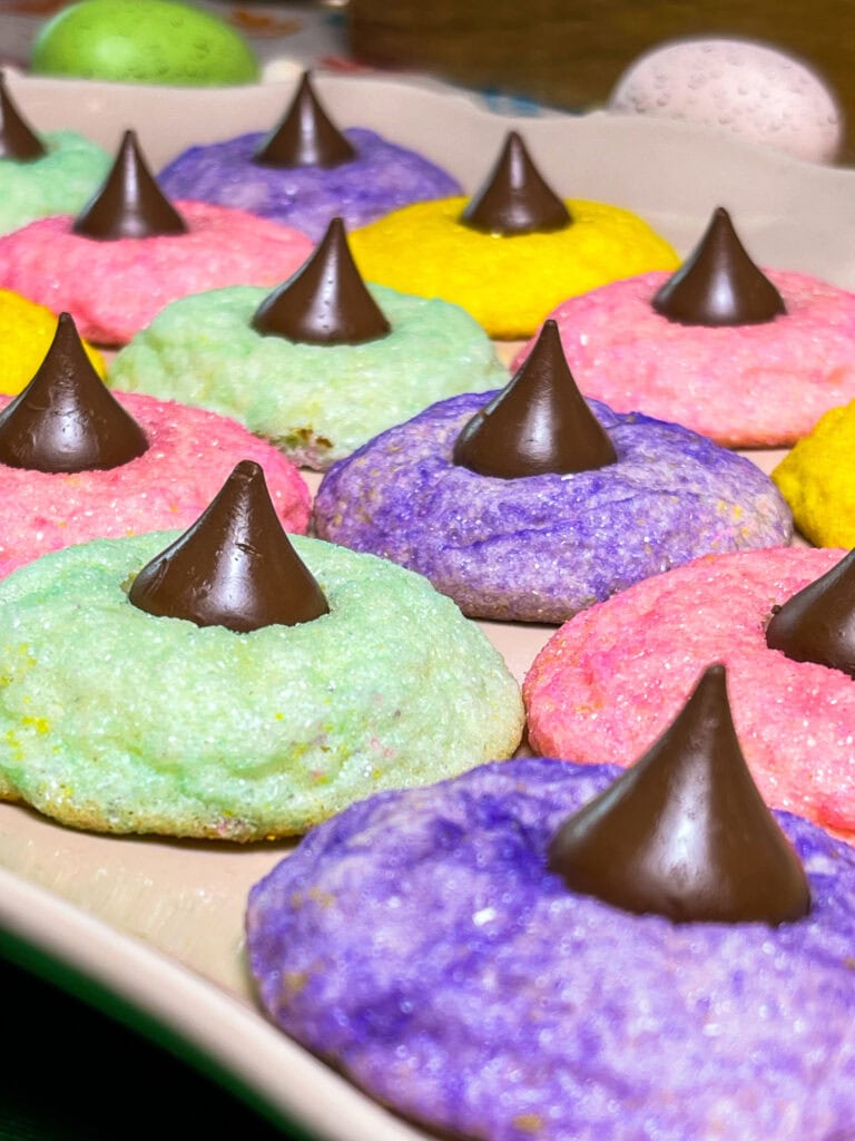 Colorful pastel sugar cookies topped with chocolate kisses are arranged on a beige tray. Perfect for your St. Patrickโs Day Treat Ideas Roundup, the cookies come in green, purple, pink, and yellowโeach with a chocolate kiss in the center.